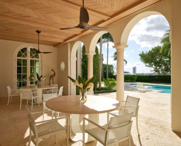 A bright, elegant covered outdoor dining terrace opening onto a swimming pool and landscaped grounds. The space features cream stucco walls, tall arched openings with classical columns, large arched glazed doors, a warm patterned timber ceiling, ceiling fans, and pale outdoor dining furniture. A city skyline is visible beyond the pool.