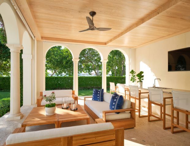 Bright covered outdoor living and dining area with a warm timber-lined ceiling, cream rendered walls, classical arched openings and columns, and views onto a manicured garden. The space includes timber-framed lounge furniture with blue cushions, a coffee table, dining table and chairs, a compact kitchenette or bar with cabinetry, and a ceiling fan. The overall design is refined, resort-like, and Mediterranean-influenced.