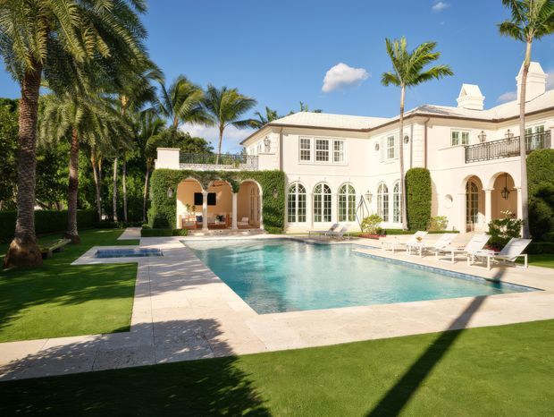 Exterior view of a grand two-storey Mediterranean-inspired residence beside a large rectangular swimming pool. The pale stucco facade features arched openings, French-style windows, decorative cornices, wrought-iron balcony railings, and clipped hedges. A manicured lawn, tall palm trees, tiled pool surrounds, and a covered outdoor loggia create a resort-like tropical setting under a clear blue sky.