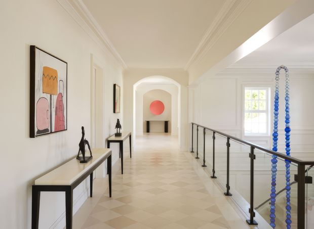 Bright, refined interior hallway with a long patterned stone or tile floor, white walls, ornate crown moulding, arched openings, framed artwork, slim console tables, and a black metal-and-glass balustrade overlooking a lower level. A striking blue bead-like vertical installation hangs beside the stair opening.