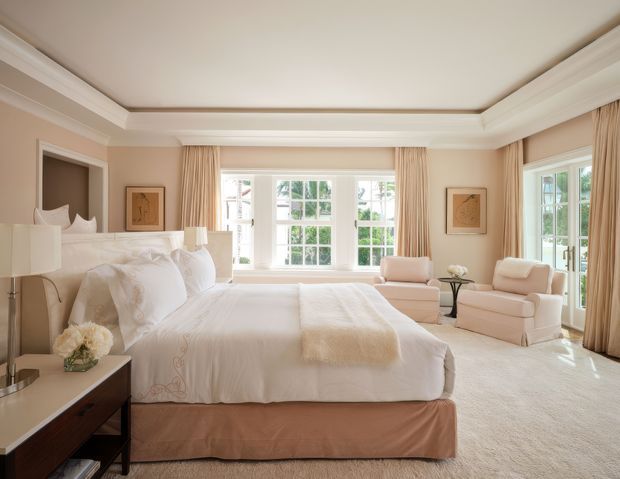 A bright, elegant bedroom with a large upholstered bed dressed in white linens, matching cream armchairs and ottoman, floor-to-ceiling beige curtains, and large divided-light windows. The room features traditional architectural detailing including crown moulding, paneled ceiling trim, and white window casings, with soft neutral furnishings and artwork.