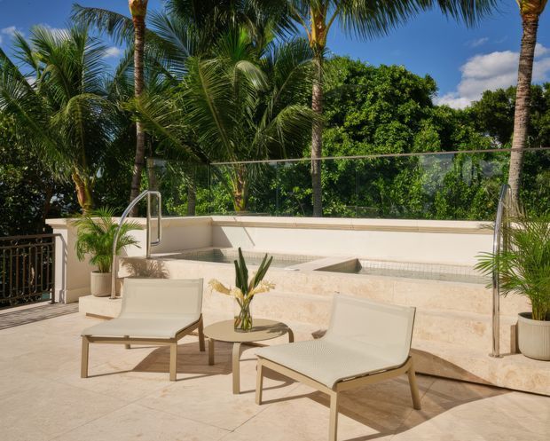 A sunlit tropical pool terrace with a shallow rectangular pool or spa, pale stone paving and retaining walls, frameless glass balustrading, palm trees, and two contemporary outdoor lounge chairs around a small table.