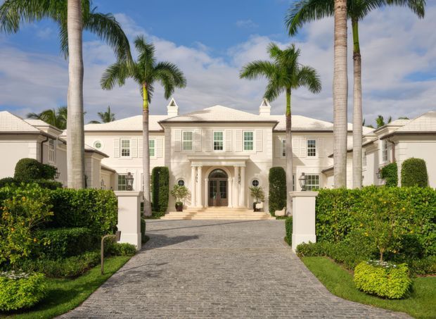 Front exterior view of a grand symmetrical pale stucco residence with a formal central entrance, classical columns, pedimented details, tiled low-pitched roofs, manicured hedges, palm trees, and a broad cobblestone driveway. The bright tropical setting gives the property a resort-like character.