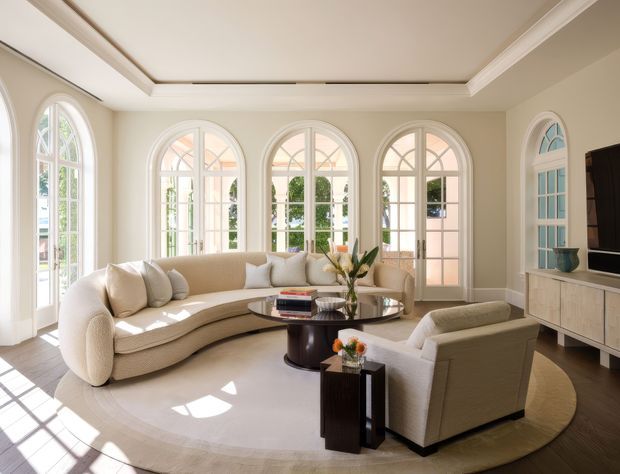 A bright, refined living room with a large curved cream sectional sofa, sculptural armchair, round coffee table, and neutral area rug. The room features tall arched multi-pane windows and doors, white traditional trim, pale walls, a recessed coffered ceiling detail, and a light built-in media console. Sunlight creates geometric shadows across the floor and furnishings.