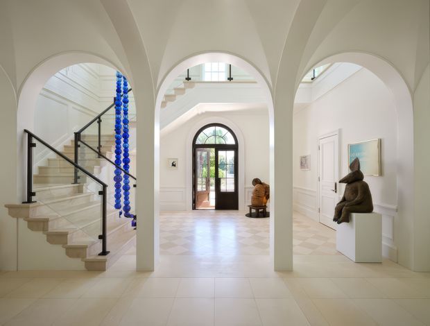 A bright, elegant residential entry foyer viewed through three large arched openings. The space features a sweeping staircase with a black metal balustrade, white decorative wall panelling, arched doorways and glazing, a pale checkerboard-style floor, and sculptural artwork displayed on a pedestal. A blue hanging decorative installation adds a vivid accent near the stairs.
