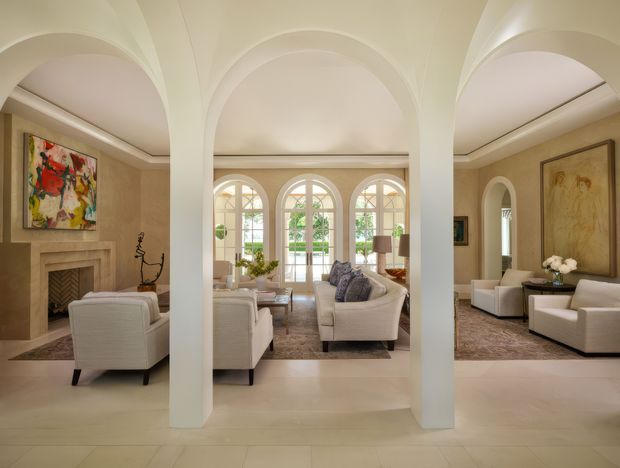 Elegant, spacious formal living room with a symmetrical arrangement of white arched columns and vaulted arch openings. The room features cream stone or tile flooring, beige plaster walls, a carved fireplace, tall arched French doors and windows, upholstered ivory sofas and armchairs, subdued artwork, and a refined neutral palette. The composition shows connected seating areas and abundant natural light.