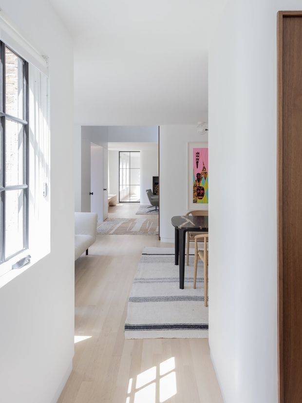 A bright, narrow interior hallway looking through a sequence of open doorways into a contemporary living and dining area. White walls and ceilings, pale timber flooring, large black-framed windows, and abundant natural light create an airy minimalist setting. A dark dining table with light timber chairs, a striped runner rug, partial white seating, and colourful wall art are visible deeper in the home. A warm timber doorway or cabinet edge frames the right side.