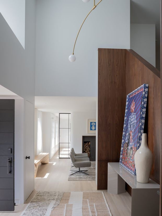 A bright, minimalist residential entryway looking into an open living area. The foreground features a dark door, pale walls, a large geometric rug, and a sculptural white pendant. A rich walnut-toned built-in timber partition and display console define the right side, with artwork, a ceramic vase, and a glimpse of a lounge, fireplace, and glazed internal door beyond.