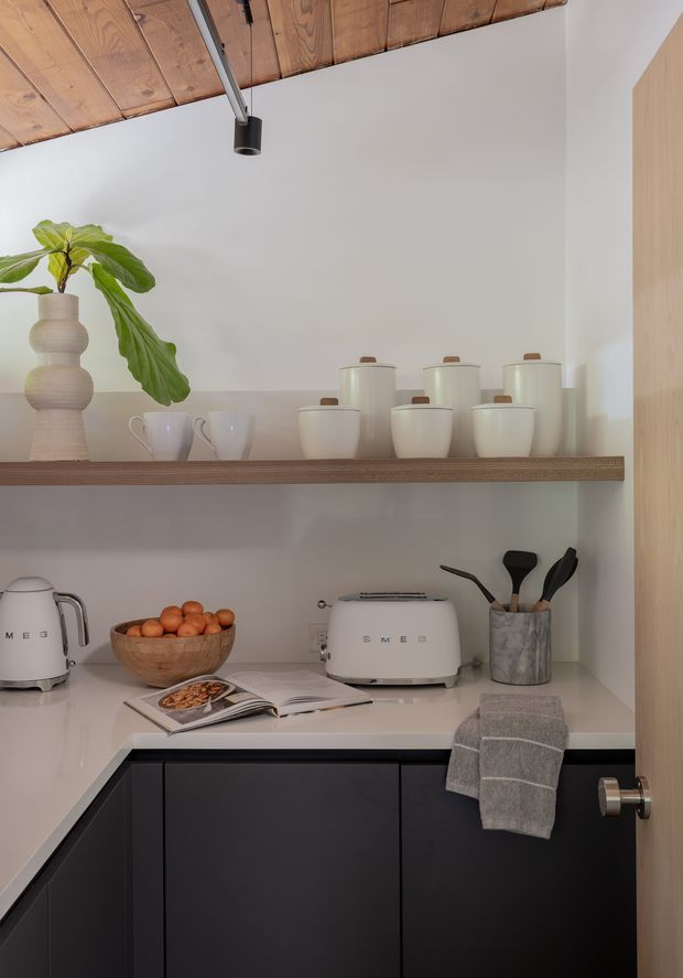 A compact, carefully styled kitchen or butler’s pantry corner with dark lower cabinetry, a light stone-look benchtop, open timber shelving, and white ceramic storage canisters and cups. A toaster, kettle, fruit bowl, utensils, cookbook, and folded tea towel sit on the benchtop. The space has white walls, a warm timber plank ceiling, black track lighting, and a tall timber panel at right.