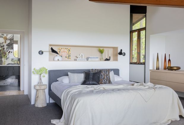 A contemporary bedroom with a low upholstered grey bed, layered neutral bedding and decorative cushions. A recessed wall niche behind the bed displays artwork and accessories, flanked by small black wall sconces. The room includes a pale built-in dresser with sculptural vases, a tall narrow window, soft grey carpet, and a warm timber ceiling beam.