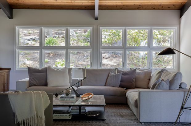 Bright, calm living room with a large L-shaped upholstered sectional, glass coffee table, floor lamp, and soft neutral furnishings. A broad bank of divided windows fills the rear wall, while exposed timber ceiling boards and dark structural beams add warmth and architectural character.