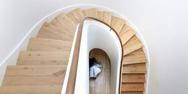 Top-down view of a sculptural curved staircase with pale timber treads arranged in a sweeping spiral around a central void. The staircase has smooth white curved balustrade walls and a slim warm timber handrail, creating a clean, contemporary architectural feature.