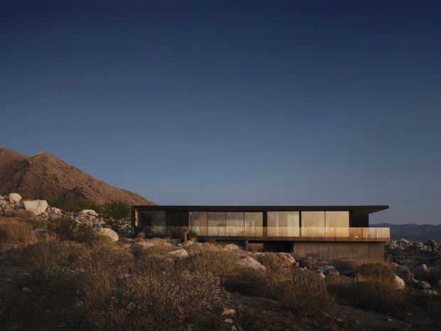 A low, elongated modern residence set into a rugged rocky hillside. The building has a strong horizontal flat roof, extensive floor-to-ceiling glazing, dark structural framing, and warm timber-toned cladding or interior surfaces. The surrounding terrain is arid and mountainous, with sparse scrub and exposed boulders, photographed in subdued late-day light.