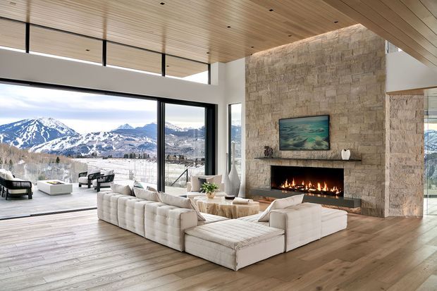 A spacious contemporary living room with a large white sectional sofa arranged around a low coffee table and a wide, linear fireplace set into a full-height natural stone feature wall. Floor-to-ceiling glazing opens to a mountain landscape and outdoor seating area. The room has a warm timber-planked ceiling and timber flooring, black-framed windows, and a high, open architectural volume.