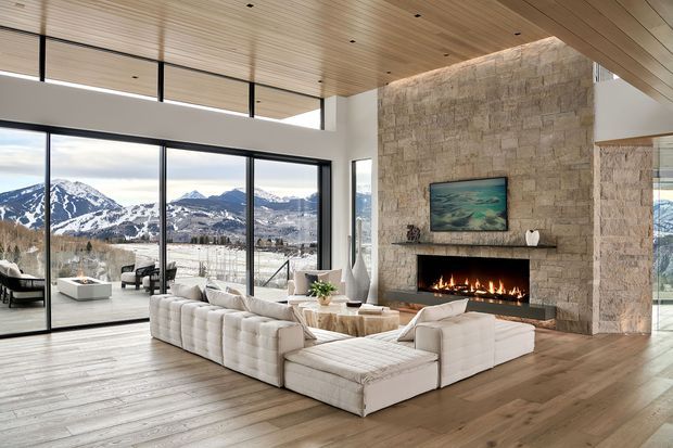 Spacious contemporary living room with a large white sectional sofa arranged around a low coffee table and a wide linear fireplace set into a full-height textured stone feature wall. Floor-to-ceiling black-framed glazing opens to expansive mountain views, while a warm timber-lined ceiling and wide timber flooring create a refined natural interior. The room has abundant daylight and a strong indoor-outdoor connection.