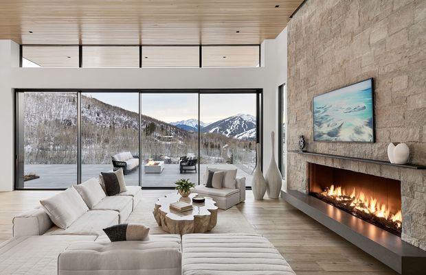 A spacious, refined living room with a large white sectional sofa and chaise, sculptural light-toned coffee table, lounge chairs, and a long linear fireplace integrated into a full-height natural stone feature wall. Floor-to-ceiling glazing opens the room to a dramatic snowy mountain landscape, while a light timber slatted ceiling and pale timber flooring create a warm, contemporary alpine atmosphere.