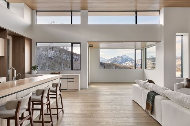 Bright open-plan contemporary interior combining a kitchen, dining and living area. The space has pale timber flooring, a long timber kitchen island with integrated cabinetry and bar seating, warm timber ceiling lining, white walls, and extensive black-framed glazing. Mountain views are visible through the windows, while a large white sectional sofa anchors the living area.