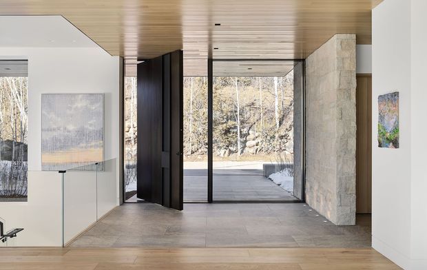 A refined modern entryway with a spacious open connection to an outdoor terrace. The interior features floor-to-ceiling glazing, large-format grey stone flooring, warm timber ceiling and flooring, slim black-framed glass doors, a dark pivot door, and a substantial pale stone feature wall. Minimal furnishings and artwork create a calm, gallery-like atmosphere.