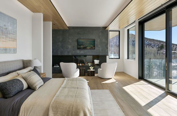 A bright contemporary bedroom with a sitting area, upholstered bed, and expansive glazed doors opening to a balcony or terrace. The room features warm timber ceiling and flooring, a dark textured feature wall with integrated joinery, white upholstered lounge chairs, and mountain scenery visible outside.