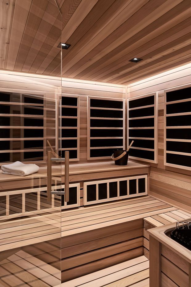 A warm, wood-lined sauna interior with tiered bench seating, horizontal timber wall slats, a timber-planked ceiling, recessed lighting, glass door, and a sauna bucket and ladle. The space has dark inset backrests or window-like panels between the timber framing and a compact sauna heater visible in the foreground.