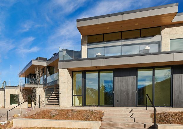 Contemporary multi-level residential facade with strong horizontal lines, flat roof forms, broad glazing, projecting balconies, and a mix of pale masonry, dark vertical cladding, grey metal panels, and warm timber soffits. Exterior stairs, terraces, balustrades, and landscaped raised beds are visible under a bright blue sky.