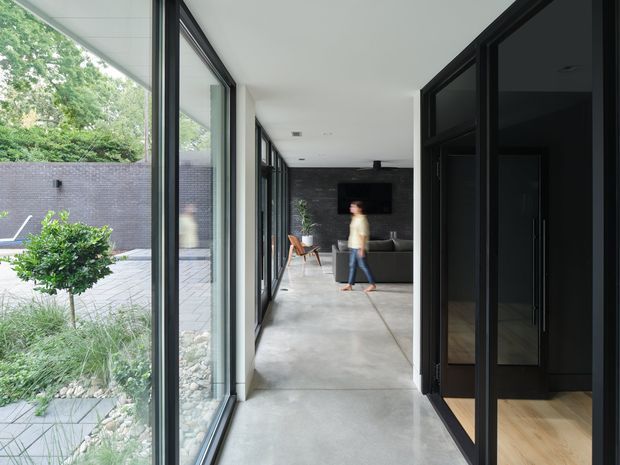 A long, contemporary interior passage connects a garden courtyard to a dark-toned living area. Full-height glazed panels line the left side, opening views to landscaped planting, paving and a rendered boundary wall. The corridor has a smooth concrete floor, white ceiling with recessed downlights, and dark-framed glazing and joinery on the right. A black feature wall, television and sofa are visible at the far end, with warm timber flooring in an adjoining room.