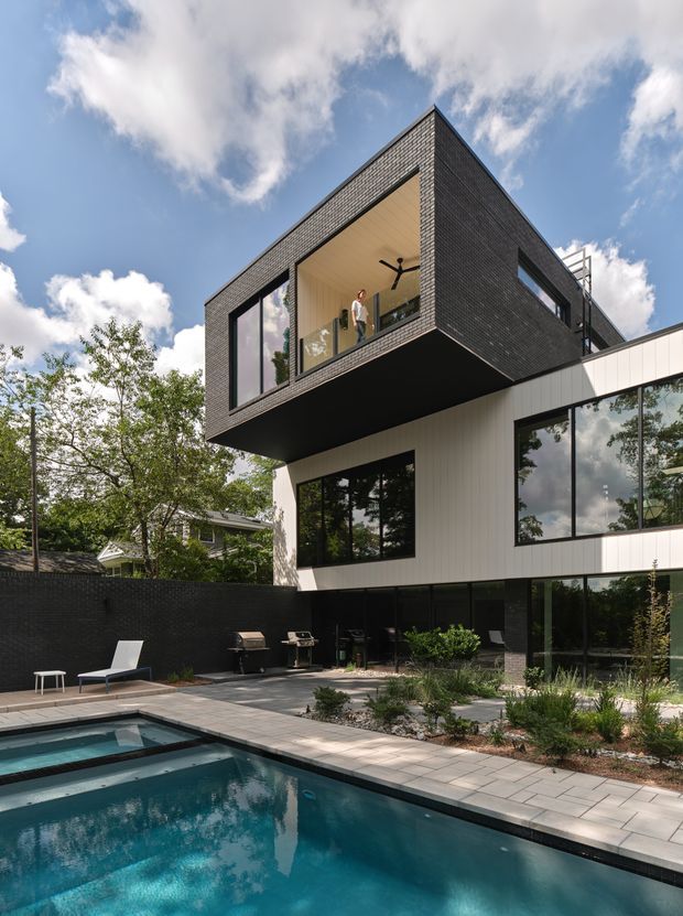 Exterior architectural photograph of a contemporary multi-level residence with a dramatic cantilevered upper volume clad in dark brick, contrasting pale vertical cladding, and extensive black-framed glazing. The house overlooks a landscaped courtyard with a swimming pool, paved terrace, outdoor seating and barbecue area. A projecting double-height glazed opening reveals a warm timber-lined interior ceiling and ceiling fan. Mature trees and blue sky surround the property.