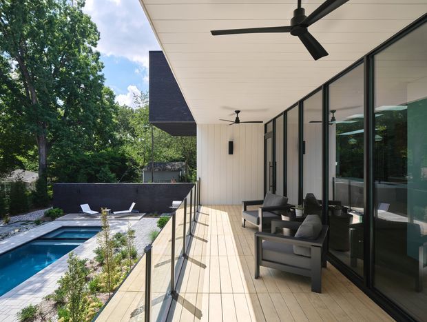 A contemporary covered outdoor terrace overlooking a swimming pool and landscaped garden. The terrace has pale timber-look decking, black-framed floor-to-ceiling glazing, slim glass balustrades, a white-lined ceiling with multiple black ceiling fans, and dark outdoor lounge furniture. The setting is leafy and residential, with strong indoor-outdoor connection and abundant daylight.