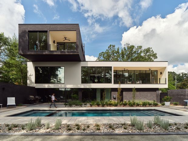Contemporary multi-level residence with bold cantilevered upper volumes, dark brick or masonry cladding, crisp light-colored horizontal walls, and extensive black-framed glazing. The house overlooks a rectangular swimming pool with paved surrounds and minimalist landscaped planting. Bright daytime exterior view with a blue sky and trees in the background.