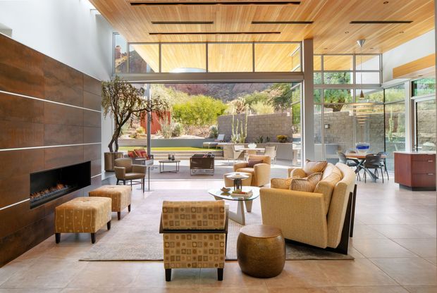 A bright, spacious contemporary living room with floor-to-ceiling glazing opening toward a landscaped courtyard. The room features a dramatic dark wood feature wall with a built-in fireplace, a warm timber slatted ceiling with recessed linear lighting, pale large-format flooring, and a generous seating arrangement with sofas, armchairs, ottomans, and a central coffee table. A dining area and kitchen cabinetry are visible to the right, with a strong indoor-outdoor connection and high clerestory glazing.