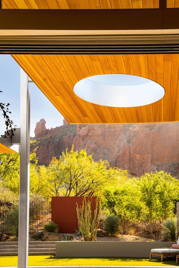 A dramatic architectural exterior view framed by a deep timber-lined soffit with a large oval skylight opening. Slim metal structural posts and expansive glazing overlook a landscaped terrace with drought-tolerant planting, a bright yellow-green outdoor surface, and a rocky red desert cliff beyond. The composition emphasizes indoor-outdoor connection, strong geometric forms, warm timber, and a striking natural setting.