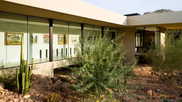 Single-storey modern courtyard-like building viewed from an arid landscaped garden. Broad floor-to-ceiling glazing with slim dark frames opens onto a sheltered interior, beneath a low projecting flat roof and pale rendered walls. Desert planting, cacti, gravel, rocks and scrub surround the building, with artwork and interior circulation visible through the windows.