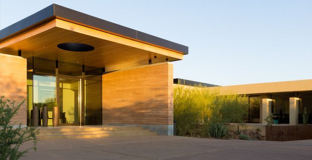 A low-slung contemporary building exterior with a prominent sheltered entry pavilion. The entrance features warm timber soffit and wall cladding, expansive glass doors, dark metal roof edging, recessed circular lighting, and broad concrete steps. A connected single-storey wing extends to the right behind landscaped planting, with a paved forecourt and desert-style vegetation.