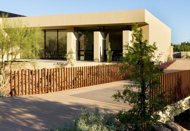 Exterior view of a low, modern single-storey residence with a flat roof, pale rendered walls, extensive dark-framed glazing, and a recessed covered entry or outdoor living zone supported by square columns. A broad paved driveway or forecourt leads toward the house, bordered by a vertical timber slat fence and drought-tolerant landscaping with sculptural cacti and shrubs.