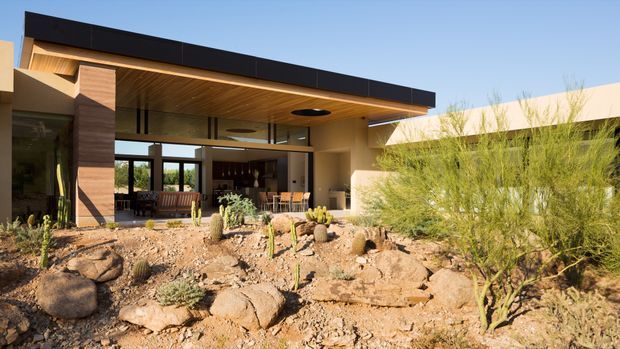 Exterior view of a contemporary single-storey residence with a broad flat roof and deep covered outdoor living area. The facade combines dark cladding, warm timber soffit lining, rendered walls, structural columns, and extensive floor-to-ceiling glazing. A dry desert-style landscape with rocks, gravel, cacti, and drought-tolerant shrubs occupies the foreground, while an open-plan interior is visible through the glazing.