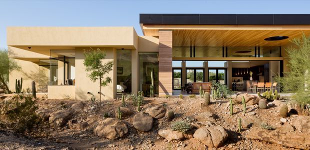 Wide exterior view of a low-slung contemporary residence set within a rocky, drought-tolerant landscape. The home has strong rectilinear volumes, warm earth-toned rendered walls, extensive floor-to-ceiling glazing, a deep covered outdoor living area, dark flat roof edging, and a timber-lined soffit. The covered space appears to connect to an open-plan living or dining area, with desert planting and exposed boulders in the foreground.