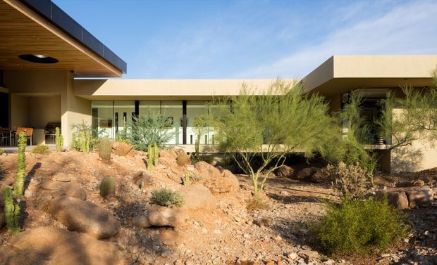 Single-storey contemporary residential building viewed from a landscaped front or courtyard area. The architecture features broad flat roof planes, projecting eaves, dark metal fascia, extensive glazed openings, and warm neutral rendered walls. The foreground is a deliberately arid xeriscape with exposed rocks, gravel, desert shrubs, and cacti. A timber-lined soffit is visible beneath the left roof overhang.
