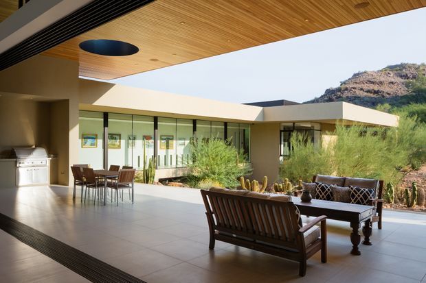 A covered outdoor living and dining terrace adjoining a low, modern home. The space has a broad tiled floor, timber-lined soffit with integrated linear ventilation and a circular recessed light, glazed walls, an outdoor barbecue, dining table and chairs, and a lounge arrangement facing a landscaped rocky hillside. The architecture features strong horizontal planes, expansive glazing, and an indoor-outdoor connection.