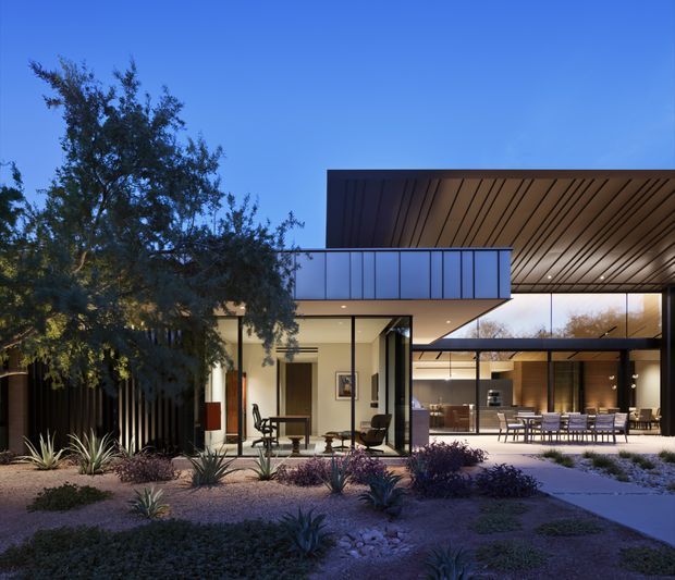 Twilight exterior view of a contemporary single-storey residence with strong indoor-outdoor connections. The house features extensive floor-to-ceiling glazing, a broad flat roof with a warm timber-lined soffit, dark slender window frames, and a covered outdoor entertaining area with a long dining table. The landscaped foreground uses gravel, rocks, agave-like plants, and low shrubs, while mature trees frame the building.