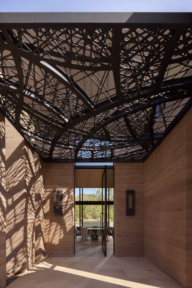 A tall, narrow architectural passage or entry court with warm sandstone walls, a dark sculptural perforated canopy overhead, and strong patterned shadows cast across the walls and floor. Glazed double doors open toward a furnished interior and landscaped view beyond. The composition emphasizes dramatic overhead structure, natural light, and geometric shadow play.