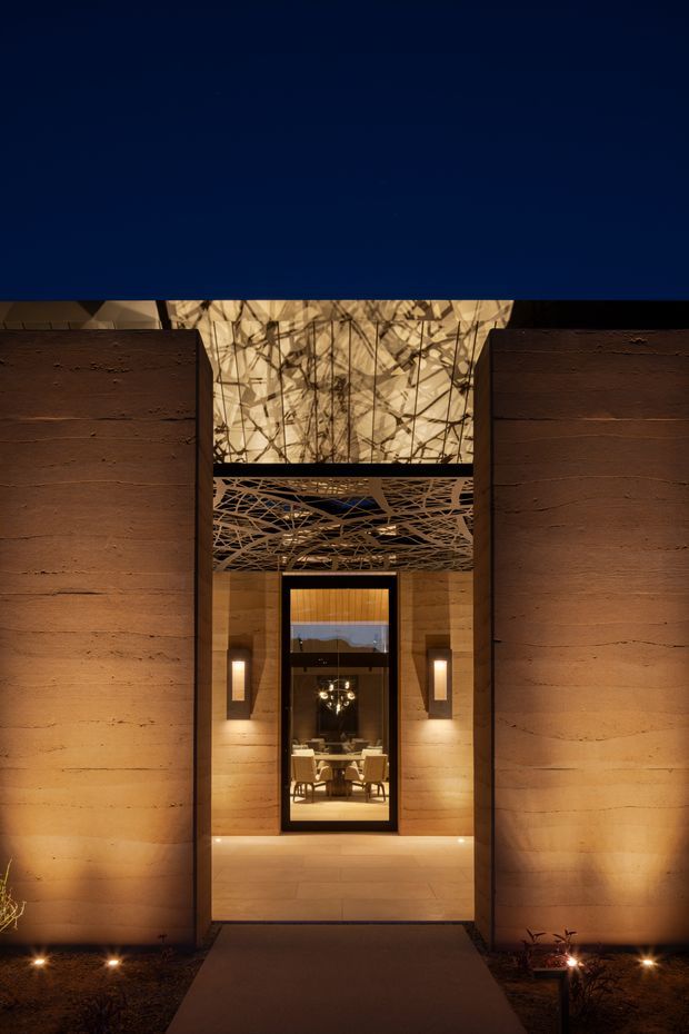 Dramatic dusk view of a monumental modern entrance framed by tall, warm earth-toned textured walls. A recessed illuminated passage leads to a glazed front door, with a striking organic branch-like patterned ceiling and translucent overhead feature panel. Subtle landscape lighting lines the dark planted approach, while a glimpse of a refined interior dining area is visible through the doorway.