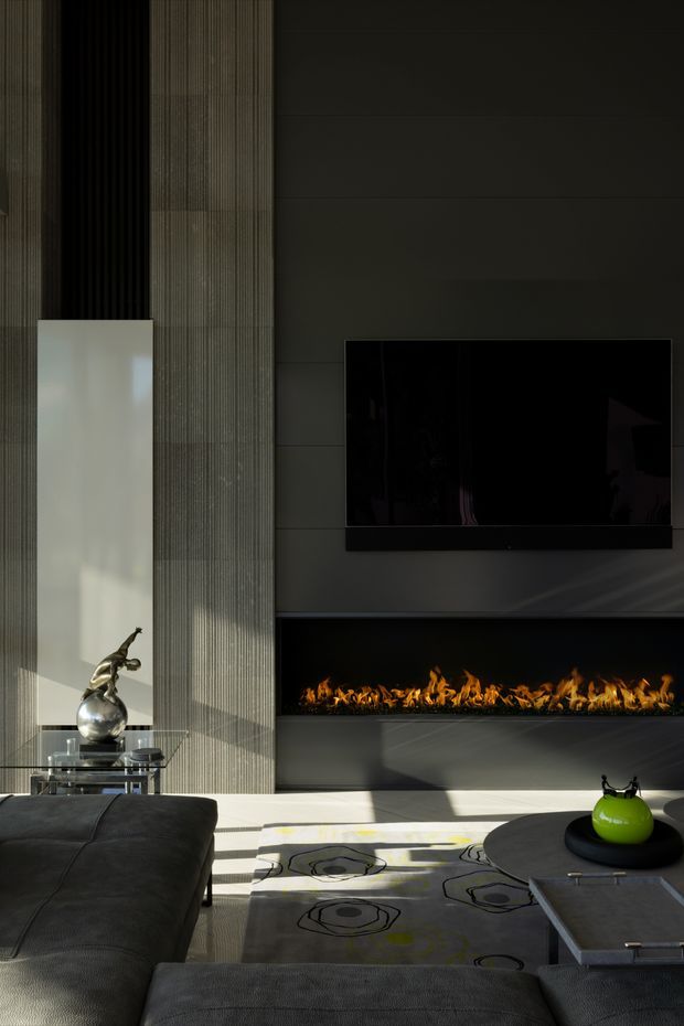 A dark, sophisticated contemporary living room centred on a long linear gas fireplace with a wall-mounted television above. The feature wall combines dark horizontal panels with a tall vertically ribbed textured section and a large pale inset panel. A charcoal sectional sofa, glass side table with sculptural decor, patterned rug, and low dark coffee table complete the room. Strong sunlight creates dramatic diagonal shadows across the floor and furniture.