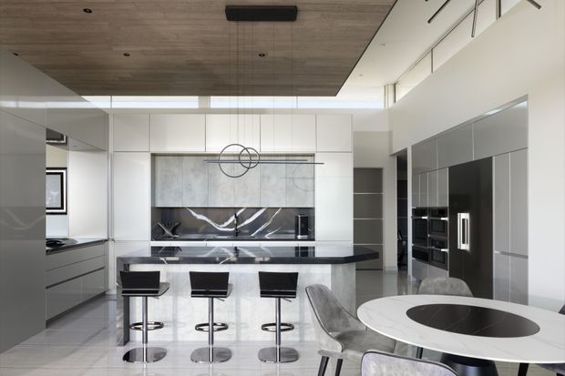 A bright, spacious contemporary kitchen and dining area with a large central island, three black bar stools, extensive white and grey cabinetry, integrated appliances, dramatic dark marble-look splashback, and a round dining table in the foreground. The room features a warm timber ceiling panel, polished light flooring, high clerestory glazing, and minimalist pendant lighting.