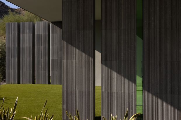 Close-up exterior view of a contemporary building with tall, vertically ribbed dark facade screens or cladding panels. Deep roof overhangs and strong sunlight create sharp geometric shadows across the facade, while narrow green-tinted glazing is visible between the panels. A manicured lawn and planted foreground frame the structure, with a rocky vegetated hillside in the background.