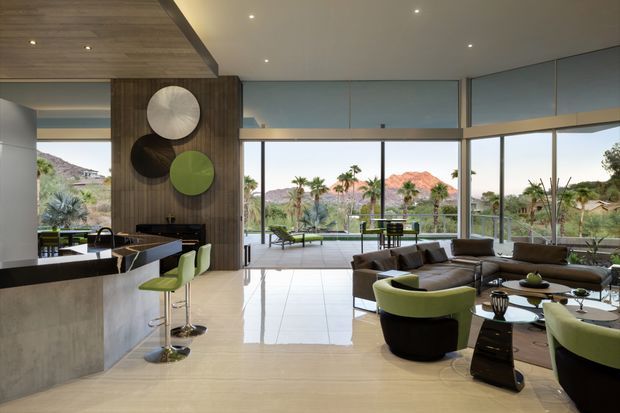 A spacious, open-plan contemporary living and kitchen area with floor-to-ceiling glazing overlooking a landscaped setting and rocky hills. The room features a large stone island with lime-green bar stools, dark wood wall panelling, a streamlined sectional seating arrangement, polished light flooring, recessed ceiling lighting, and an adjacent outdoor terrace visible through the glazing.