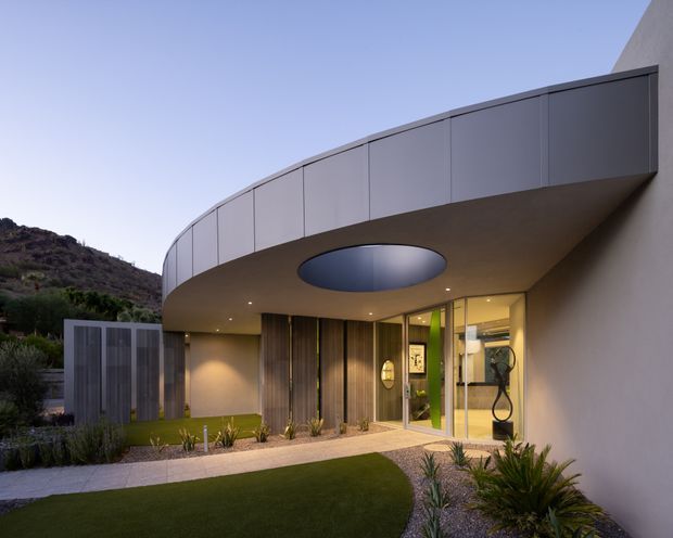 Exterior view of a contemporary, curved-roof residential entrance set into a sloping landscaped site. The building features smooth light-coloured walls, a dark grey curved fascia, recessed lighting, floor-to-ceiling glazed entry doors, vertical timber-toned screening, and a prominent oval skylight in the sheltered canopy. A paved walkway and planted landscaping lead to the entry.