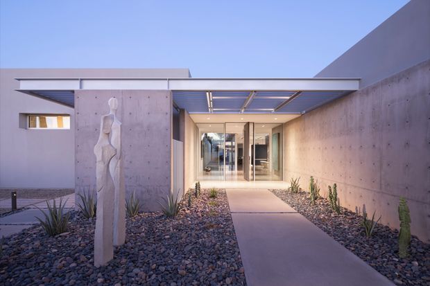 Exterior view of a minimalist modern residential entry, framed by smooth concrete walls and a flat projecting roof canopy. A straight paved walkway crosses a dry gravel landscape with sparse architectural planting and a sculptural vertical feature, leading to a glazed entrance. The image is photographed at dusk with warm interior lighting visible through the doors.