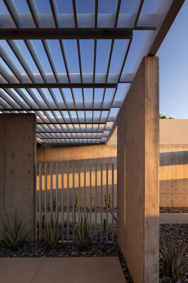 A narrow contemporary courtyard or covered outdoor passage framed by raw concrete walls and substantial timber posts. A slatted pergola roof with translucent panels creates strong linear shadows and filtered daylight overhead. The ground is covered with dark decorative gravel and drought-tolerant agave-like plants, with a concrete walkway in the foreground and a slim metal security or privacy fence across the middle.
