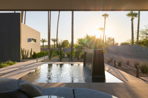 A refined modern outdoor courtyard viewed from a shaded covered lounge, with a rectangular reflecting pool and central dark sculptural water feature. Pale rendered walls, large-format paved surfaces, desert planting, cacti, and tall palm trees create a warm, minimalist resort-like setting in low golden sunlight.