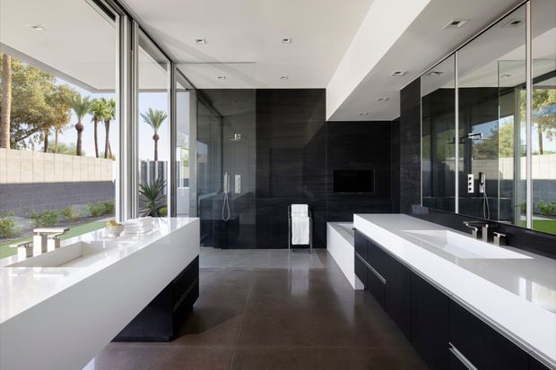 Contemporary open-plan kitchen and living space with a dramatic black feature wall, integrated fireplace or television niche, and extensive floor-to-ceiling glazing. A large white island and parallel white benchtop create a bright contrast against dark fixed cabinetry and large-format grey flooring. The glazing opens visually to a landscaped outdoor area with palms.